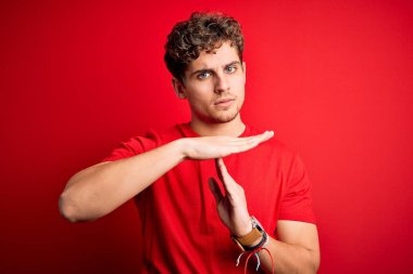 Young blond handsome man with curly hair wearing casual t-shirt over red background Doing time out gesture with hands, frustrated and serious face
