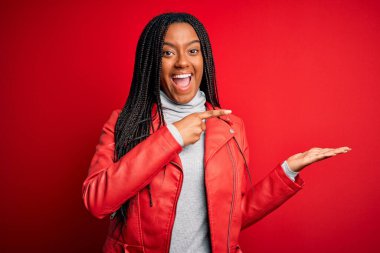 Genç Afrikalı Amerikalı kadın, kırmızı izole edilmiş arka planda havalı bir deri ceket giyiyor. Kameraya hayret ediyor ve gülümsüyor..