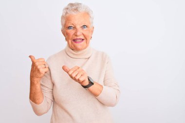 Senior grey-haired woman wearing turtleneck sweater standing over isolated white background Pointing to the back behind with hand and thumbs up, smiling confident