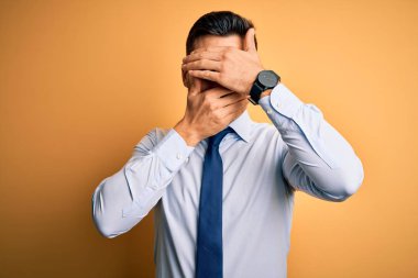 Sarı arka plan üzerinde duran kravat ve gözlük takan genç yakışıklı işadamı elleri ve ağızları kapalı, şaşırmış ve şok olmuş. Duygularını gizliyor.