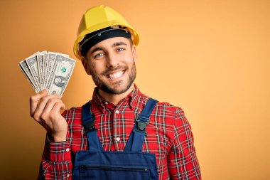 Güvenlik kaskı takan genç müteahhit, sarı arka plan üzerinde ödeme olarak dolar tutuyor. Mutlu bir yüzü var, gülümsüyor ve kendine güvenen bir gülümsemeyle dişlerini gösteriyor.
