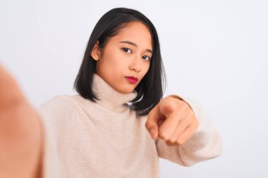 Balıkçı yaka kazak giyen genç Çinli bir kadın izole edilmiş beyaz arka plan üzerinde selfie çekerek kamerayı işaret ediyor ve sen, el işareti, ön taraftan pozitif ve kendinden emin bir hareket.