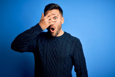 Sıradan bir süveter giyen genç bir adam izole edilmiş mavi arka planda dikiliyor. Şok olmuş bir şekilde yüzünü ve gözlerini kapıyor. Utanmış bir ifadeyle parmaklarının arasından bakıyor..