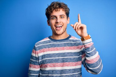 Young blond handsome man with curly hair wearing striped sweater over blue background pointing finger up with successful idea. Exited and happy. Number one.