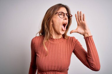 Young beautiful brunette woman wearing casual sweater and glasses over white background shouting and screaming loud to side with hand on mouth. Communication concept.