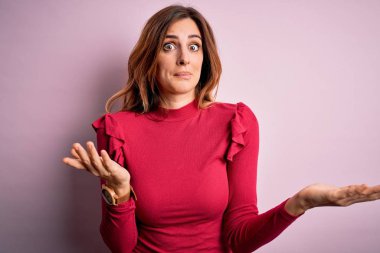 Young beautiful brunette woman wearing casual sweater stnding over pink background clueless and confused expression with arms and hands raised. Doubt concept.