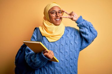 Young African American student woman wearing muslim hijab and backpack holding book Doing peace symbol with fingers over face, smiling cheerful showing victory