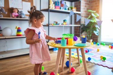 Anaokulunun masasında çatal bıçak ve yemek oyuncaklarıyla oynayan genç ve güzel bir çocuk.