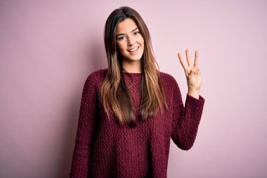 İzole edilmiş pembe arka plan üzerine sıradan süveter giyen genç ve güzel bir kız üç numaralı parmaklarıyla işaret ederken kendinden emin ve mutlu bir şekilde gülümsüyor..