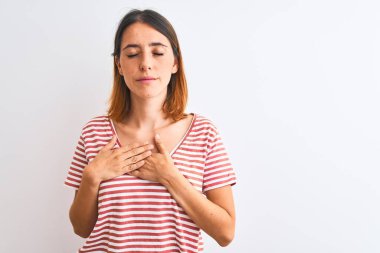 Güzel kızıl saçlı kadın izole edilmiş arka planda sıradan kırmızı tişörtler giyiyor. Elleri göğsünde, gözleri kapalı ve yüzünde minnet dolu jestlerle gülümsüyor. Sağlık kavramı.