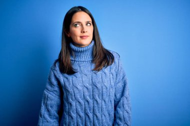 Mavi gözlü genç esmer kadın balıkçı yaka kazak giyiyor gülümsüyor ve yan tarafa bakıyor ve düşünüyor..