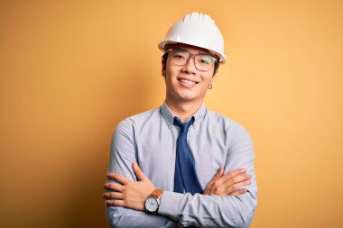 Young handsome chinese architect man wearing safety helmet and tie over yellow background happy face smiling with crossed arms looking at the camera. Positive person.