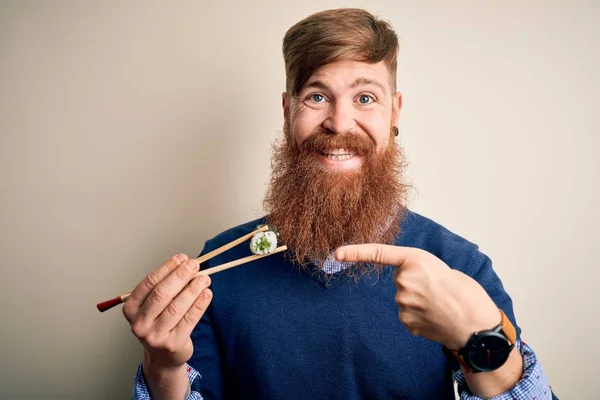 Redhead Irish man with beard eating green maki sushi using chopsticks over yellow background very happy pointing with hand and finger