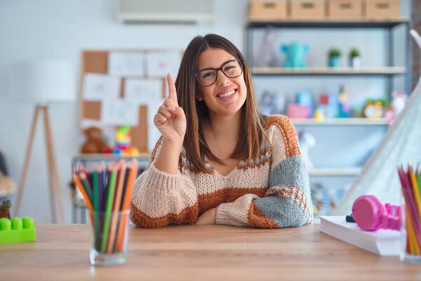 Young beautiful teacher woman wearing sweater and glasses sitting on desk at kindergarten showing and pointing up with finger number one while smiling confident and happy.