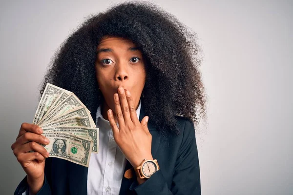 Young african american business woman with afro hair holding cash dollars banknotes cover mouth with hand shocked with shame for mistake, expression of fear, scared in silence, secret concept