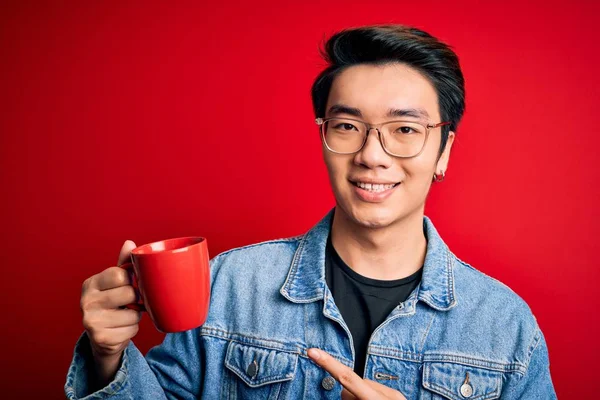 Young handsome chinese man drinking cup of coffee over isolated red background very happy pointing with hand and finger