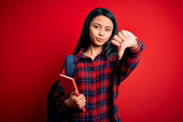 Young beautiful chinese student woman holding book standing over isolated pink background with angry face, negative sign showing dislike with thumbs down, rejection concept