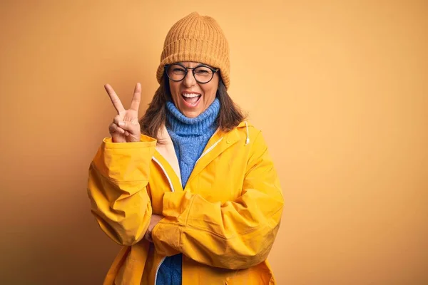 Middle age woman wearing yellow raincoat and winter hat over isolated background smiling with happy face winking at the camera doing victory sign. Number two.