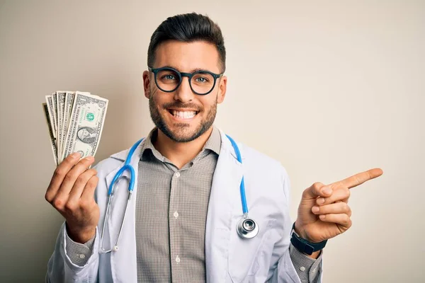 Young doctor man holding a bunch of one dollar banknotes over isolated background very happy pointing with hand and finger to the side