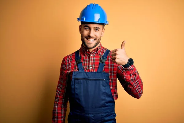 年轻的建筑工人 身穿建筑制服 头戴安全帽 头戴黄色背景 手举着快乐的大拇指 在镜头前看着成功的表情 — 图库照片