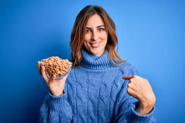 Young Beautiful Brunette Woman Holding Bowl Peanuts Blue Background Surprise — 图库照片