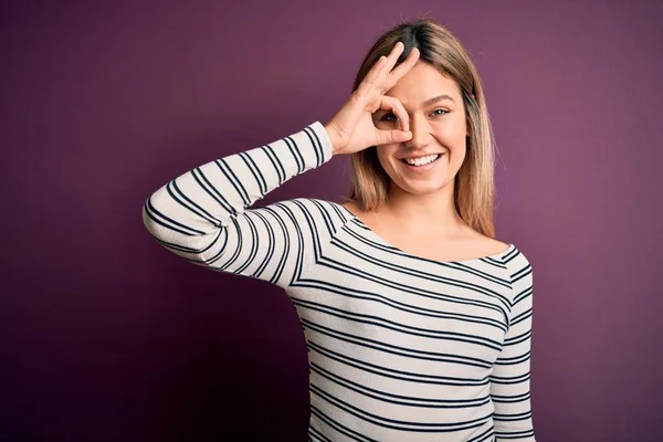 Young beautiful blonde woman wearing casual striped shirt over purple isolated background doing ok gesture with hand smiling, eye looking through fingers with happy face.