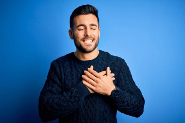 İzole edilmiş mavi arka plan üzerinde duran, elleri göğsünde, gözleri kapalı ve yüzünde minnet dolu bir jest olan genç, yakışıklı bir adam. Sağlık kavramı.