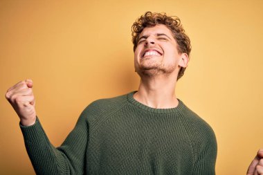 Sarı arka planda yeşil süveter giyen, kıvırcık saçlı, genç, sarışın yakışıklı bir adam. Çok mutlu ve heyecanlı. Kolları havada, gülümseyen ve başarı için çığlık atan, kazanan bir jest yapıyor. Kutlama konsepti.