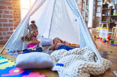 Sevimli bebekler çadırın içinde battaniyeye uzanıp gülüyorlar ve anaokulunda oyuncak bebekle oynuyorlar.