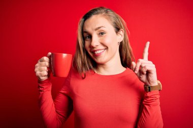 Genç, güzel, sarışın bir kadın izole edilmiş kırmızı arka planda kahve içiyor. Bir fikir ya da soru işareti ile şaşırmış, mutlu bir yüz, bir numara.