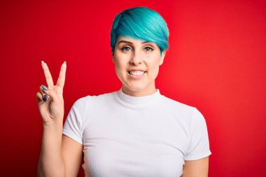 Mavi saçlı, kırmızı arka planda sıradan tişörtler giyen genç ve güzel bir kadın kameraya bakıp zafer işareti yapıyor. İki numara..