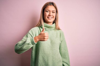 Genç, güzel, sarışın bir kadın pembe yünlü bir arka plan üzerine kazak giyip mutlu bir el hareketi yapıyor. Kameraya bakıp başarıyı gösteren ifade onaylanıyor.