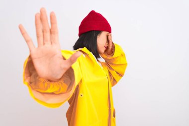 Genç, güzel Çinli bir kadın, izole edilmiş beyaz arka planda yünlü bir şapka ve yağmurluk takıyor, gözleri ellerle kaplıyor ve üzgün ve korku ifadeleriyle jest yapıyor. Utanç verici ve olumsuz bir kavram..