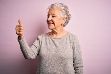 Üst düzey güzel bir kadın, günlük tişört giyip pembe arka planda dikiliyor. Gururlu görünüyor, gülümsüyor, baş parmak işareti yapıyor.