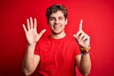 Sarışın, kıvırcık saçlı, kırmızı arka planda sıradan tişörtler giyen genç ve yakışıklı bir adam. Altıncı parmaklarını gösterirken kendinden emin ve mutlu bir şekilde gülümsüyor..