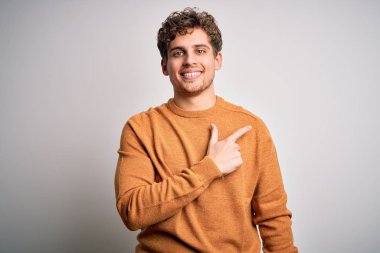 Sarışın, kıvırcık saçlı, beyaz arka planda rahat süveter giyen, yüzünde mutlu ve doğal bir ifadeyle el ve parmak gösteren bir gülümseme olan genç, yakışıklı bir adam.