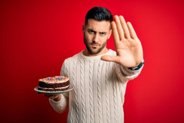Genç yakışıklı adam kırmızı izole edilmiş arka planda renkli bir doğum günü pastası tutuyor. Açık elle dur işareti yapıyor. Ciddi ve kendinden emin bir ifadeyle, savunma hareketi yapıyor.
