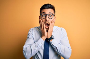 Sarı arka plan üzerinde duran kravat ve gözlük takan genç yakışıklı işadamı korkmuş ve şok olmuş, şaşırmış ve şaşırmış bir şekilde yüzü olan