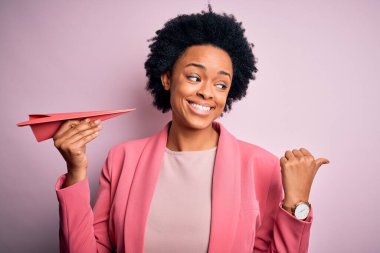 Kıvırcık saçlı, pembe kâğıt uçak tutan, gülümseyen yüzüyle baş parmağıyla yüzünü gösteren Afro-Amerikan bir kadın.