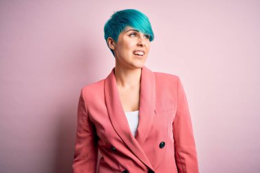 Mavi saçlı, pembe arka planda ceket giyen, yüzünde gülümseme, doğal bir ifade olan genç ve güzel bir iş kadını. Kendinden emin gülen.