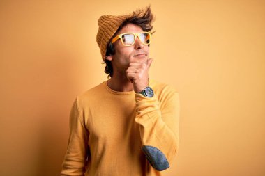 Sıradan tişört ve gözlük takan genç yakışıklı bir adam izole edilmiş sarı arka planda elleri çenesinde soru sorarken, düşünceli bir ifade takınıyor. Düşünceli bir yüzle gülümsüyor. Şüphe kavramı.