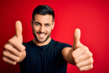 İzole edilmiş kırmızı arka plan üzerinde duran siyah tişört giyen genç yakışıklı adam el hareketlerini olumlu karşılıyor, gülümsüyor ve başarı için mutlu. Kazanan jest.
