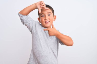 Gri günlük tişört giyen güzel çocuk izole edilmiş beyaz arka planda durup gülümsüyor ve mutlu yüzlü elleri ve parmakları olan bir çerçeve yapıyor. Yaratıcılık ve fotoğrafçılık kavramı.