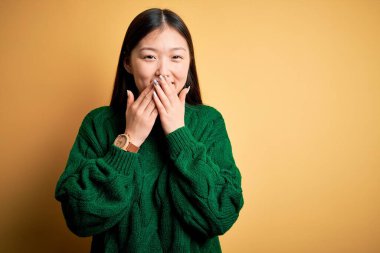 Young beautiful asian woman wearing green winter sweater over yellow isolated background laughing and embarrassed giggle covering mouth with hands, gossip and scandal concept