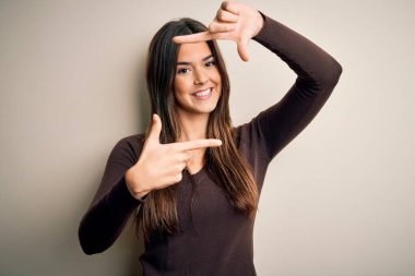 İzole edilmiş beyaz arka planda sıradan bir süveter giyen genç ve güzel bir kız gülümseyerek elleri ve parmakları gülen yüzlü bir çerçeve yapıyor. Yaratıcılık ve fotoğrafçılık kavramı.