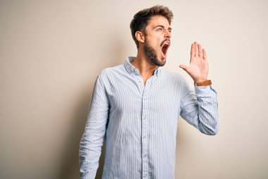 Young handsome man with beard wearing striped shirt standing over white background shouting and screaming loud to side with hand on mouth. Communication concept.