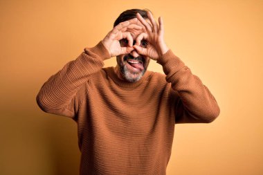 Orta yaşlı, beyaz, kahverengi süveter giyen ve gözlüklü, sarı arka planda, dürbünün dili dışarı çıkarması, gözlerin parmaklarından içeri bakması gibi hareketler yapan bir adam. Çılgın ifade.