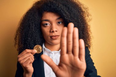 Afro saçlı, Bitcoin 'i sarı arka planda tutan, ciddi ve kendinden emin bir ifadeyle dur işareti yapan Afro-Amerikan iş kadını, savunma jesti.