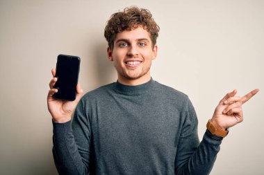 Sarışın, kıvırcık saçlı, akıllı telefon gösteren genç bir adam izole edilmiş beyaz arka planda duruyor.