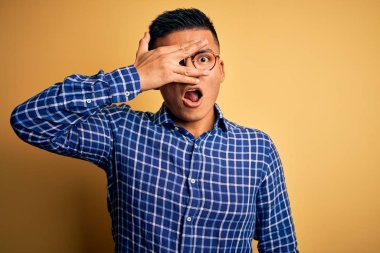 Young handsome latin man wearing casual shirt and glasses over yellow background peeking in shock covering face and eyes with hand, looking through fingers with embarrassed expression.